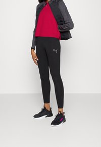 Black leggings with a white Puma logo, a dark gray puffer vest, and a red short-sleeve shirt, paired with black athletic shoes.