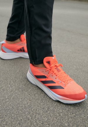 Chaussures de course en orange vif avec une tige en mesh, des rayures noires et une semelle blanche. Dotées de surfaces texturées et d'un col rembourré pour le confort.