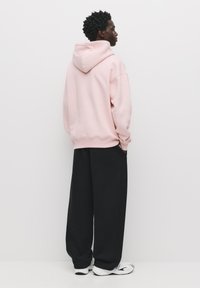 Rosa Kapuzenpullover, lässiger Schnitt, mit Kängurutasche. Kombiniert mit lockeren schwarzen Hosen und weißen Sportschuhen. Glatte Texturen sichtbar.