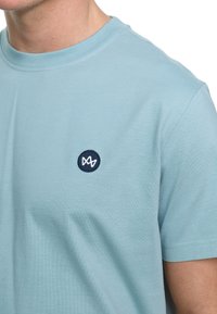 Ljusblå bomullströja med rund hals och korta ärmar. Har en rund, marinblå, broderad logotyp på vänster bröst. Mjuk textur.