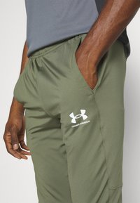 Maslinasto zelene atletske hlače s elastičnim pojasom, bočnim džepovima i bijelim Under Armour logom. Napravljene su od izdržljive, glatke tkanine.