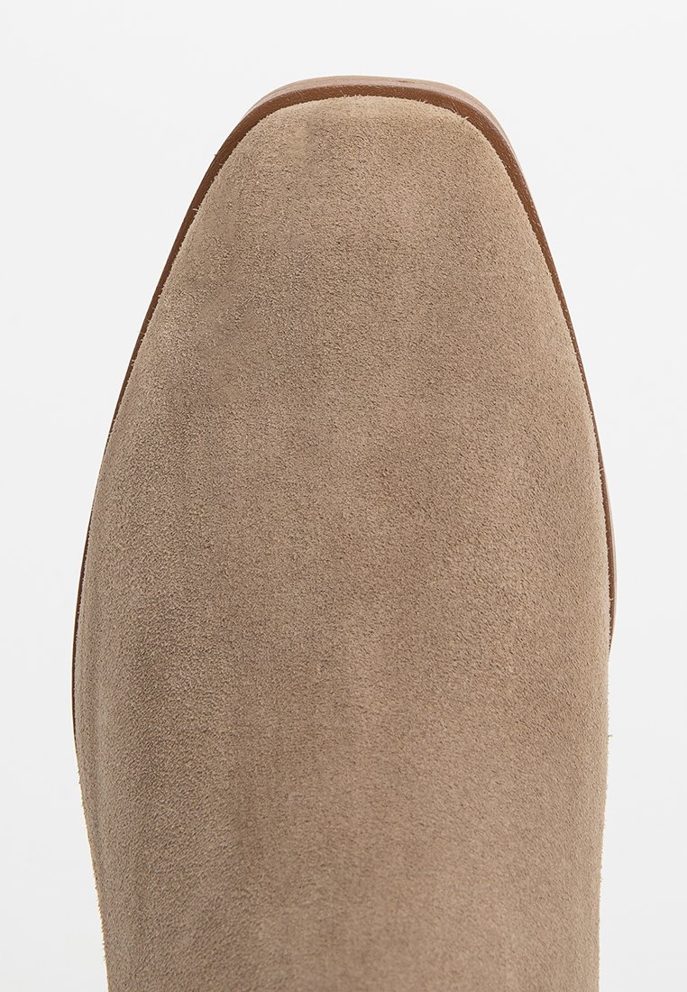 Gros plan sur l'avant d'une chaussure en suède beige avec un bout carré et un bord de semelle marron, présentée sur un fond blanc.