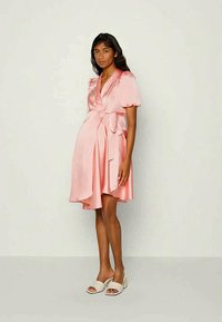 Vestido envolvente de cetim rosa com mangas bufantes, detalhe de laço na cintura e bainha assimétrica. Modelo combina com sandálias de salto aberto brancas.