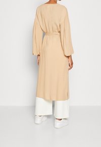 Beżowa długa sukienka z bufkowymi rękawami i wiązaniem w talii, zestawiona z białymi spodniami o szerokich nogawkach i białymi trampkami. Gładka faktura materiału.