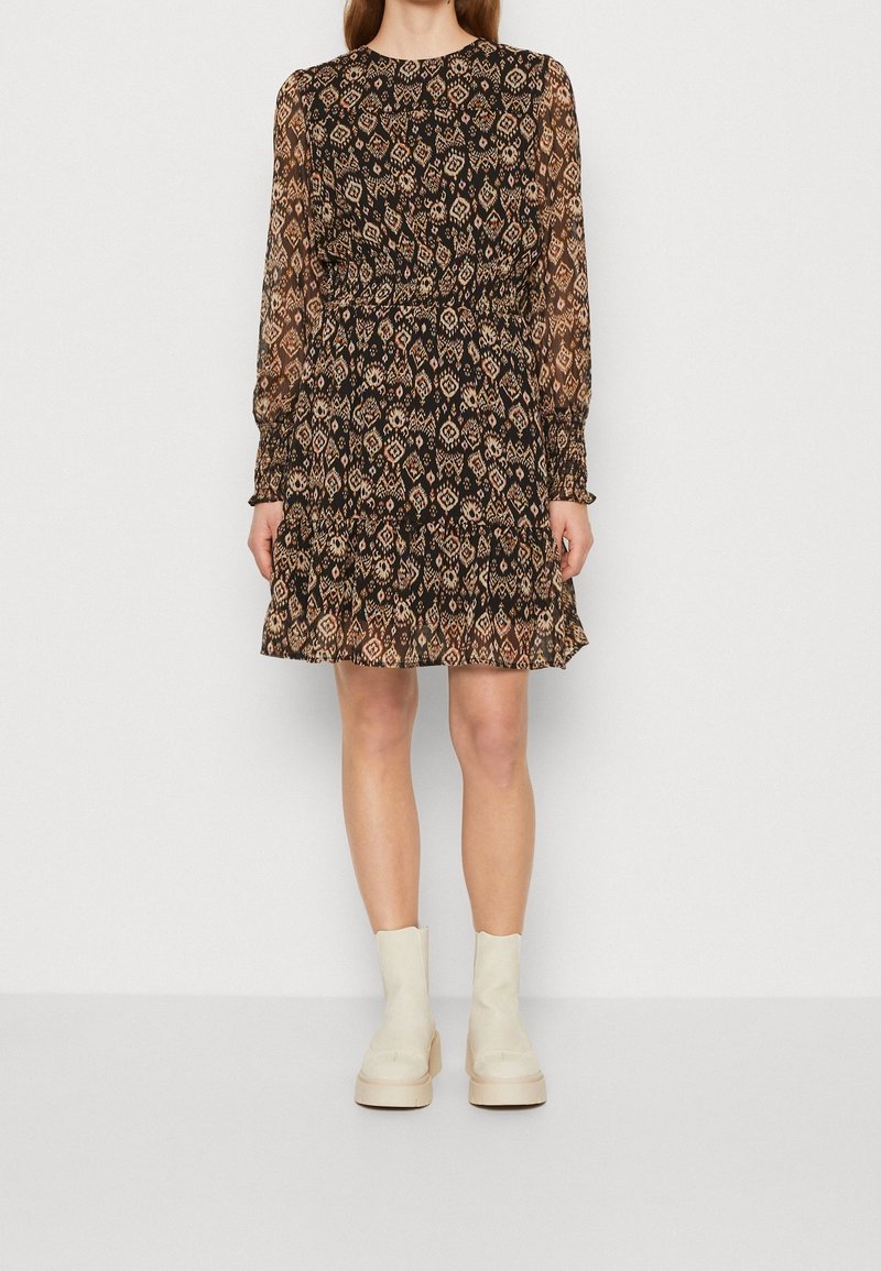 Femme portant une robe noire à motifs, manches longues, longueur genou, et des bottines blanches, debout devant un fond clair uni.