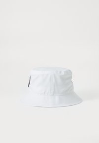 Chapeau seau blanc en tissu léger, doté d'une couronne ronde et d'un large bord inclinable vers le bas. Comprend un patch logo sur le côté.