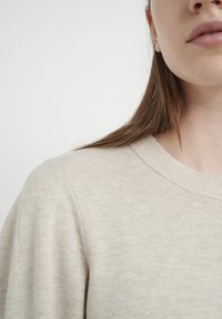 Šviesiai smėlinės spalvos megztinis su juostuotu apykakle. Minkšta tekstūra ir laisvas siluetas, fiksuojantis artimą pečių ir apykaklės vaizdą.