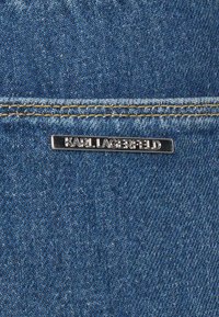 Tela de mezclilla en azul profundo con una textura suave. Presenta una etiqueta de metal plateado que dice "KARL LAGERFELD" unida a la superficie.