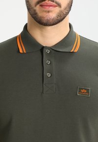 Mörkgrön piké skjorta i texturerat tyg, med en krage i orange ränder, tre knappar och en orange logotyp-patch på bröstet.