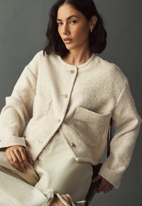 Beige textured jacket with a round neck, brass buttons, two front pockets, and long sleeves, worn over light-colored pants.