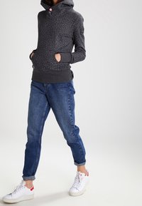 Donna con felpa a pois grigio scuro, jeans blu arrotolati e sneakers bianche, in piedi con le mani nelle tasche.