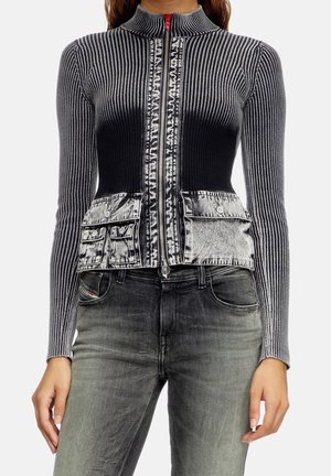 Eng anliegende schwarze und graue Rippstrickjacke mit Reißverschluss und acid-wash Vordertaschen, kombiniert mit verblassten schwarzen Jeans.