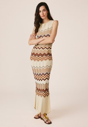 Frau mit langen dunklen Haaren steht mit verschränkten Armen, trägt ein ärmelloses, knöchellanges Kleid mit welligen braunen, beigen und cremefarbenen Mustern und gelbe Sandalen.