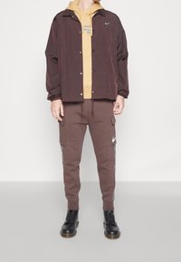 Brun nylonjacka över en gul hoodie, tillsammans med bruna cargobyxor med sidofickor och en vit logotyp, kompletterad med svarta kängor.