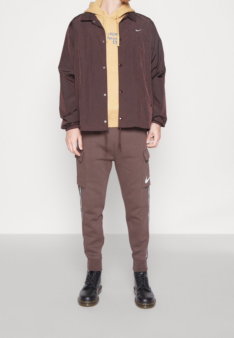 Brun nylonjacka över en gul hoodie, tillsammans med bruna cargobyxor med sidofickor och en vit logotyp, kompletterad med svarta kängor.