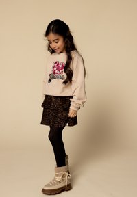 Meisje met lang donker haar, gekleed in een beige sweatshirt met een roze hart, een rok met luipaardprint, een zwarte panty en beige winterlaarzen, kijkt naar beneden.