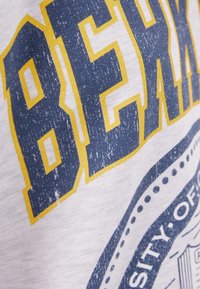 Vit T-shirt med stor blå och gul sliten text. Har ett cirkulärt emblem med en detaljerad design, delvis synlig i hörnet.