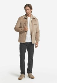 Beige kordjacka med skjortkrage, två bröstfickor, vit t-shirt under, kombinerad med mörka jeans och ljusbeiga skor.
