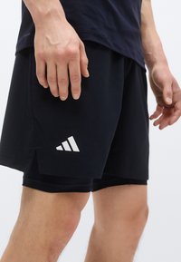 Shorts de sport noirs avec une fente latérale, présentant un logo à trois bandes blanches sur la jambe gauche. Fabriqués en tissu léger.