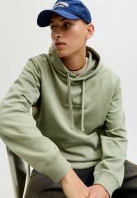 Oliva vihreä huppari, jossa on etu-tasku, nyöritettävä huppu ja ribbivaltaiset hihnat, yhdistettynä tummiin farkkuihin ja tummansinisestä lippikseen.