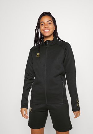 Zwarte fleece jack met rits en hoge kraag, gouden logo op de linkerborst en met rits afgesloten zakken. Draagt met bijpassende zwarte shorts.