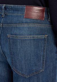 Jeans de denim azul de Trussardi con un parche de marca de cuero marrón, diseño de cinco bolsillos y detalles de costura en color naranja.