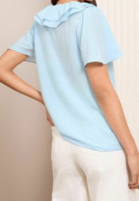 Blouse bleu clair à manches courtes avec un col à volants et un tissu lisse. Le dos présente une coupe décontractée et un léger éclat. Pantalon blanc visible en dessous.
