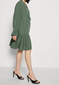 Vestido verde, de longitud a la rodilla, con mangas largas y un dobladillo con volantes. Presenta un lazo con borlas en el escote. Llevado con zapatos de tacón alto negros y de punta abierta.