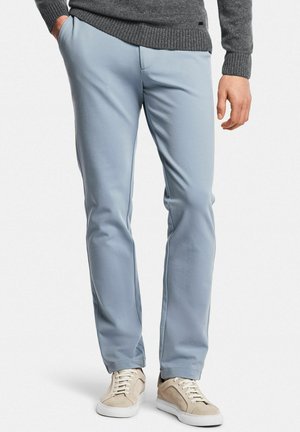 Uomo che indossa pantaloni azzurri dritti, maglione grigio e sneakers beige, in piedi con una mano in tasca.