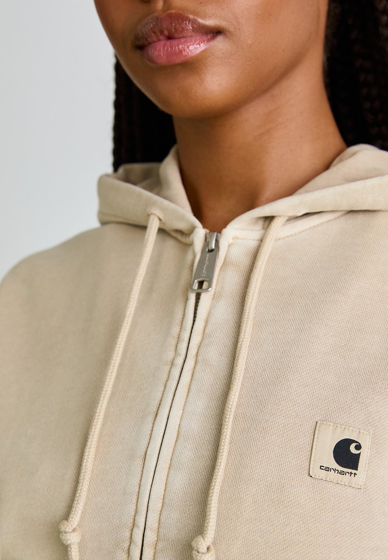 Sweat à capuche beige zippé avec cordon de serrage, en tissu texturé, et un petit patch logo sur la poitrine. Équipé d'un zip en métal argenté.