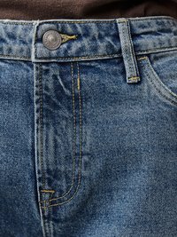 Tuvplāns zilām džinsa biksēm ar metāla pogu, dzeltenu šuvi un jostas cilpu virs priekšējā kabatas pret brūna auduma fonu.