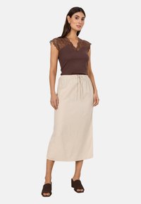 Braunes geripptes Oberteil mit Spitzenkurzärmel, kombiniert mit einem hellbeigen Midirock mit Kordelzugbund. Getragen mit dunklen Slippern.