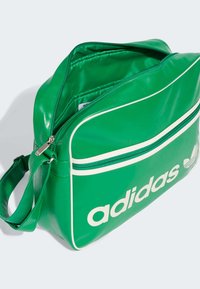 Grønn syntetisk lærveske med hvite detaljer, med en glidelåslukking hovedrom og frontlomme. Stort "adidas"-logo som vises tydelig.