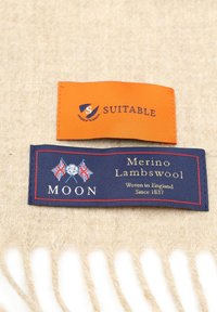 Oranje en marineblauwe labels op een lichtbeige stof met witte franjes. De labels tonen merknamen en materiaaldetails: Merino lamswol.