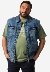 Denimväst i ljusblått med tvättad textur, försedd med två bröstfickor, metalldekor och öppen front, bärs över en grön T-shirt.
