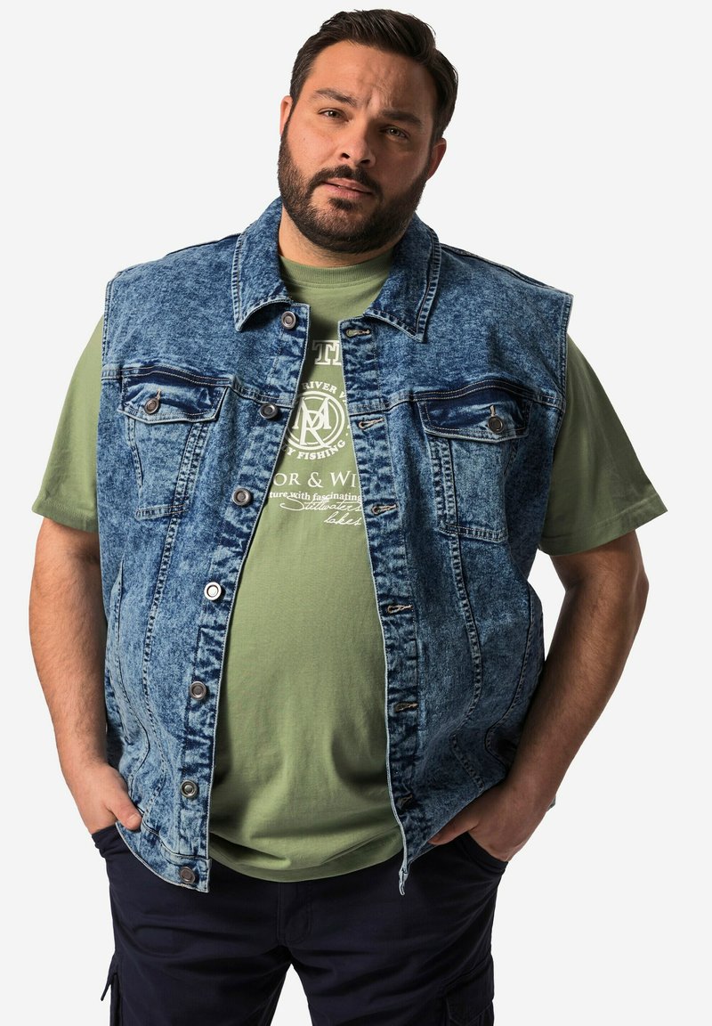 Denimväst i ljusblått med tvättad textur, försedd med två bröstfickor, metalldekor och öppen front, bärs över en grön T-shirt.