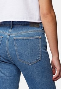 Jeans de mezclilla azul con un diseño ajustado, que cuentan con dos bolsillos traseros y una etiqueta negra en la cintura. Costuras distintivas visibles.