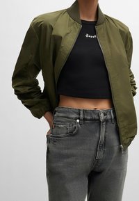 Olivi vihreä pommilentäjän takki, jossa on raidallinen kaulus, vetoketjukiinnitys ja joustavat hihansuut, yhdistetty mustan lyhyen paidan ja harmaiden farkkujen kanssa.