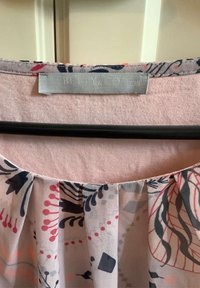 Top de tela ligera y con estampado, base rosa, con diseños florales y abstractos en azul marino, coral y gris. Presenta pliegues y una etiqueta gris.