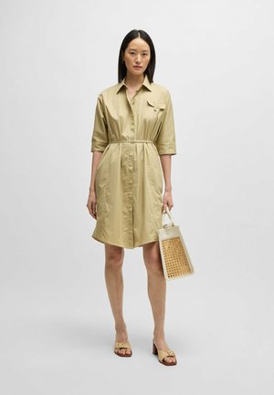 Khaki shirt dress with short sleeves, a belted waist, and a pocket. Worn with beige heeled sandals and carrying a textured white bag.
