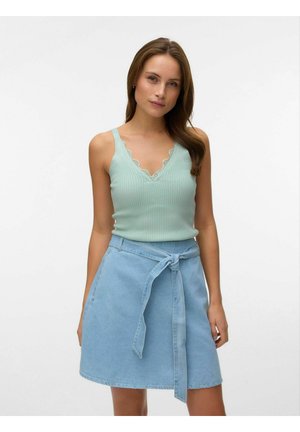 Ženska, oblečena v svetlo modro rebrasto majico brez rokavov z čipkastim robom in visokoločnato denim krilo, zavezano v pasu, stoji pred enobarvnim ozadjem.