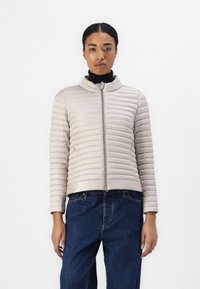Beige boblejakke med horisontal søm, høy krage og frontglidelås. Båret over en svart topp, sammen med mørkeblå jeans.