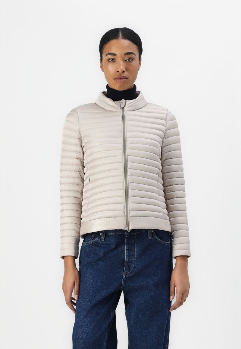 Beige boblejakke med horisontal søm, høy krage og frontglidelås. Båret over en svart topp, sammen med mørkeblå jeans.