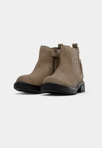 Une paire de bottines en suède taupe avec des semelles noires, fermeture éclair latérale et panneau élastique avec des perforations en forme d'étoile, présentée sur un fond blanc.