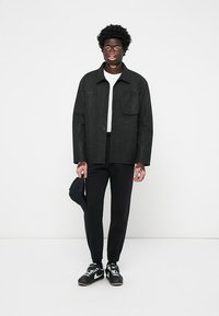 Sort tekstureret jakke med en lomme, hvid skjorte under, sorte bukser og sorte sneakers, der står foran en hvid baggrund.