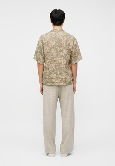 Άνδρας που είναι στραμμένος πλάτη, φοράει μπεζ κοντομάνικο πουκάμισο με λουλουδένιο σχέδιο, ανοιχτό μπεζ παντελόνι με φαρδιά μπατζάκια και λευκά αθλητικά παπούτσια, σε λευκό φόντο.