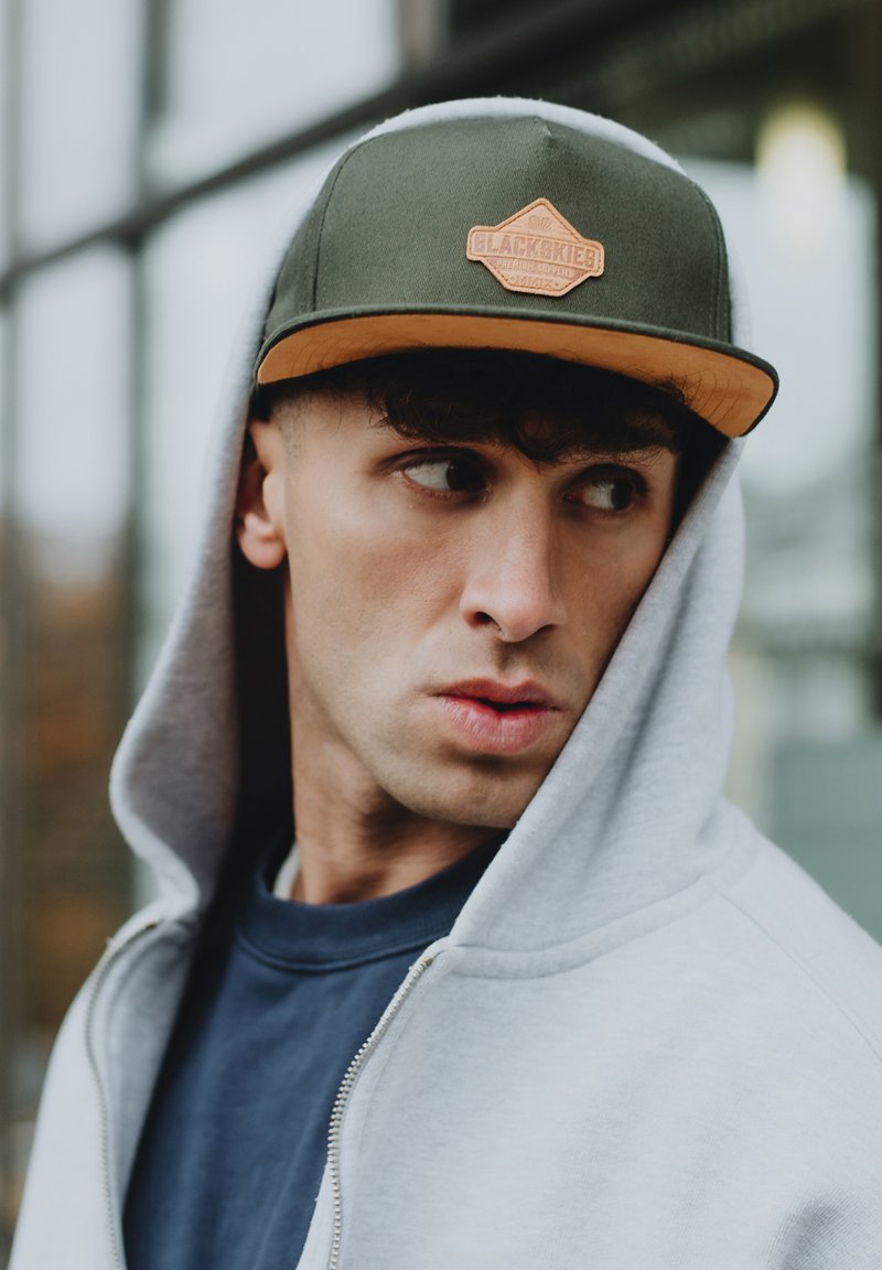 Cappello verde con logo arancione, felpa grigia con zip, e camicia blu navy. Capelli ricci e posa girata contro uno sfondo urbano sfocato.