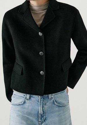 Veste noire cintrée avec trois boutons, deux poches avant, portée sur un pull à col roulé beige et un jean en denim bleu clair.