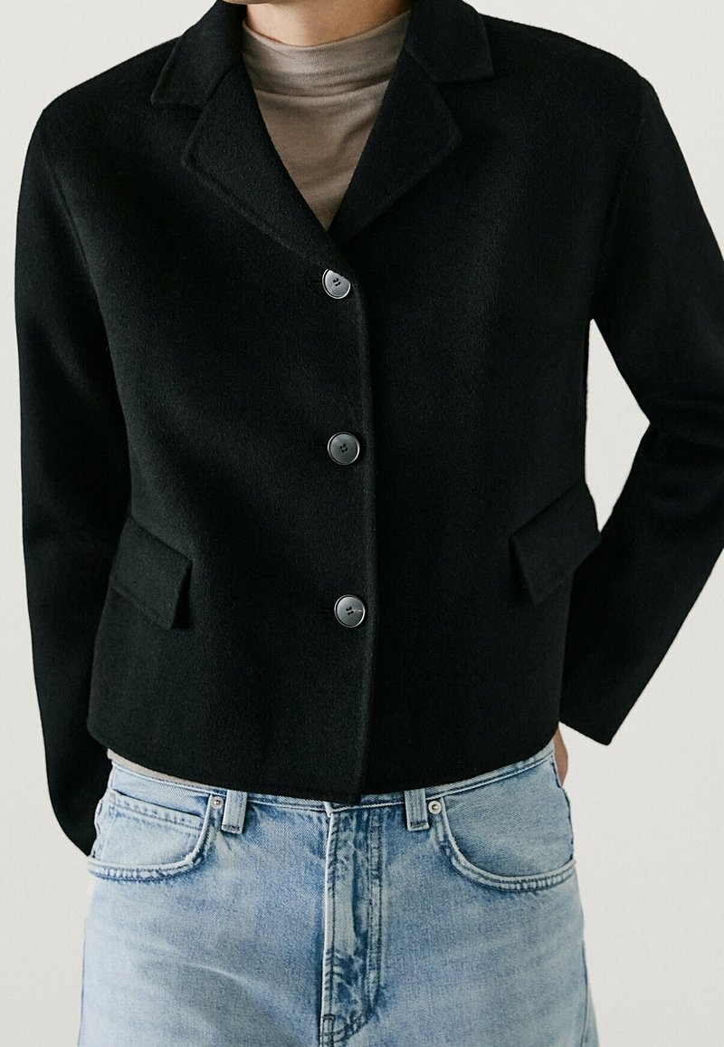 Veste noire cintrée avec trois boutons, deux poches avant, portée sur un pull à col roulé beige et un jean en denim bleu clair.