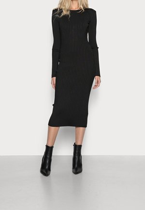 Kobieta w dopasowanej, czarnej, prążkowanej sukience midi z długim rękawem i czarnych botkach za kostkę, stojąca na jednolitym tle.